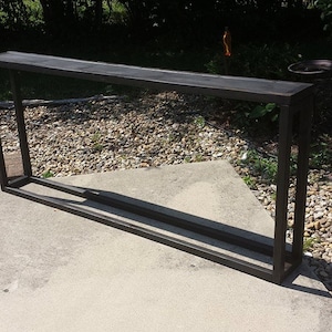 May include: A dark brown metal console table with a rectangular top and a rectangular frame. The table is approximately 60 inches long and 12 inches wide. The table is outdoors on a concrete surface.