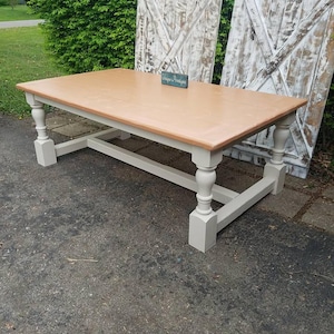 May include: A rectangular dining table with a light brown wooden top and a light gray base featuring four turned legs connected by a horizontal stretcher. A small rectangular sign with the text Unique Pruntiques sits on the table surface.