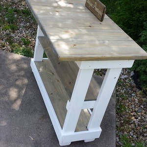 May include: White and gray wooden table with a shelf. The table has a white painted base and a gray stained top. The table is sitting on a concrete surface. A sign that says "Unique Primtiques" is sitting on the table.