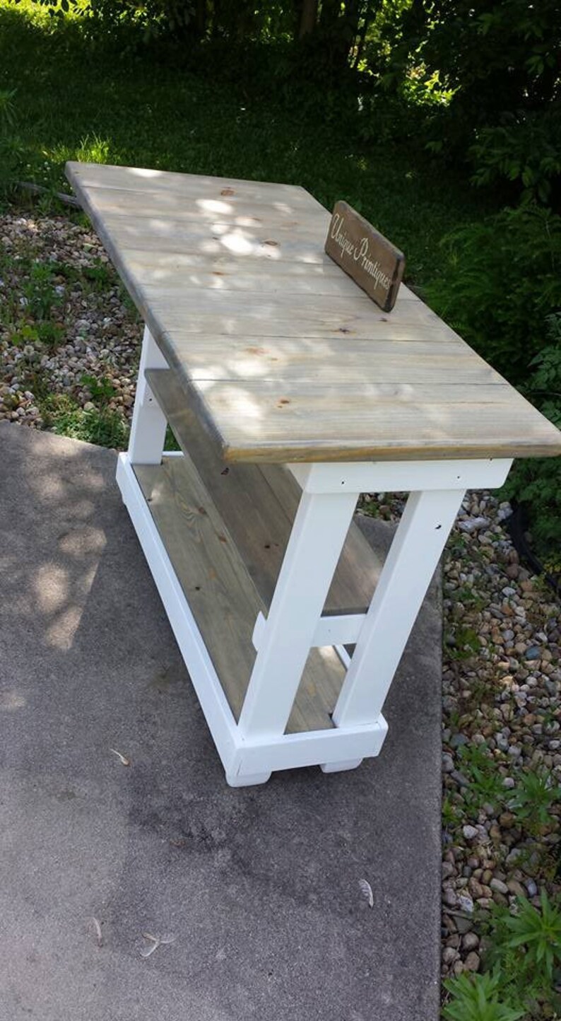 May include: White and gray wooden table with a shelf. The table has a white painted base and a gray stained top. The table is sitting on a concrete surface. A sign that says "Unique Primtiques" is sitting on the table.