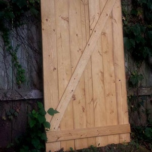 May include: A light brown wooden door with a diagonal brace and a horizontal top rail. The door is made of vertical planks and has a rustic look.