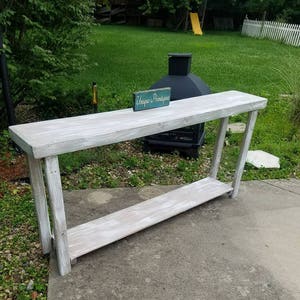 May include: Whitewashed wooden console table with a single shelf. The table is 48 inches long and 16 inches wide. The table is sitting on a concrete patio with green grass in the background. A small sign with the text "Unique Finds" is sitting on the table.