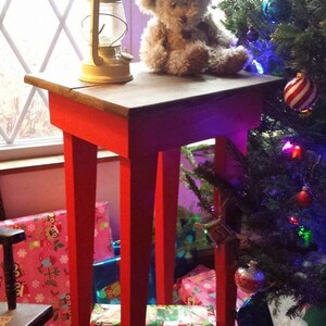 May include: A red wooden side table with a teddy bear sitting on top. The table is surrounded by wrapped gifts and a Christmas tree with ornaments.