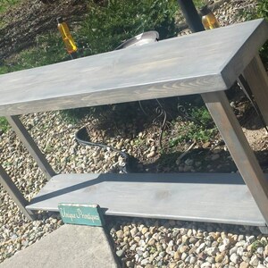 May include: A gray wooden console table with a lower shelf. The table has a rectangular top and angled legs. The wood has a visible grain and a weathered appearance. A small sign with the text "Unique Printiques" is visible.