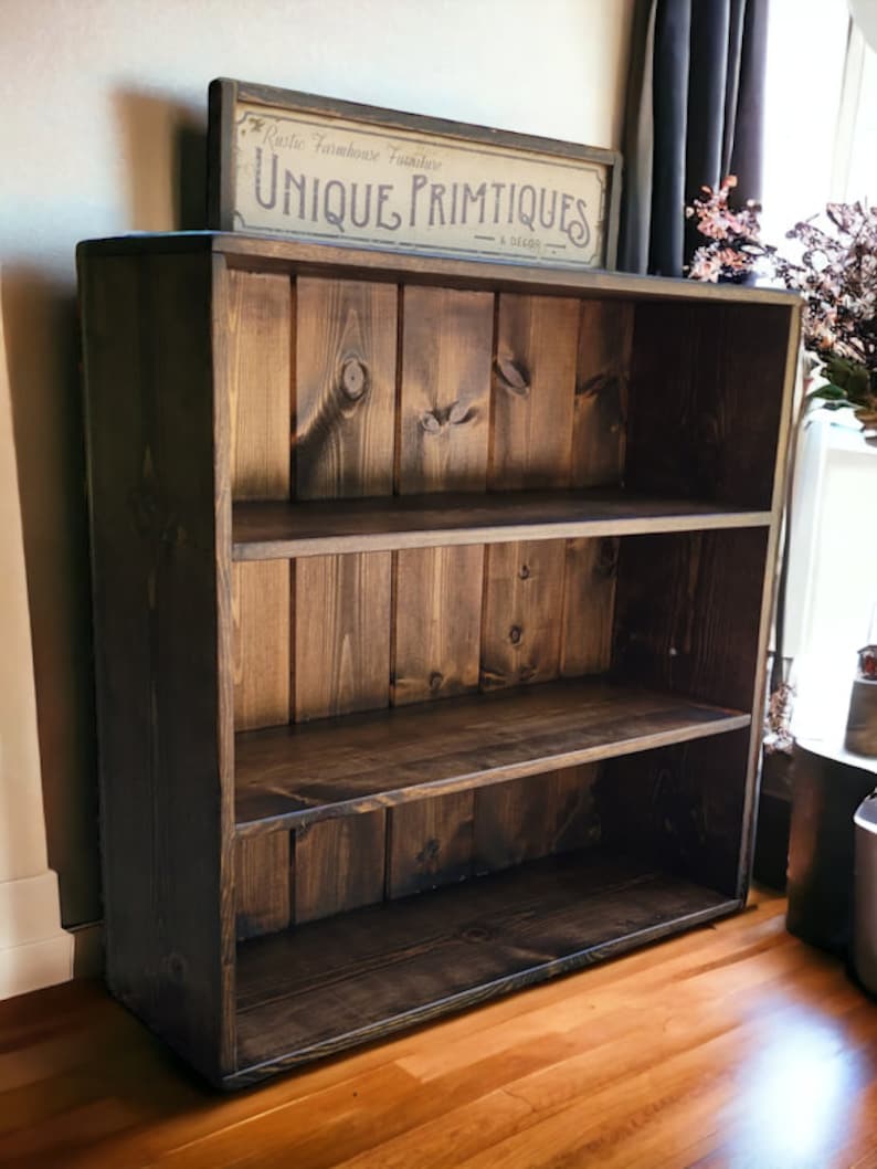 Puede incluir: Una estanter&iacute;a de madera marr&oacute;n oscuro con tres estantes. La estanter&iacute;a tiene un estilo r&uacute;stico de granja y est&aacute; hecha de madera maciza. El letrero sobre la estanter&iacute;a dice "Rustic Farmhouse Furniture Unique Primitives & Decor".