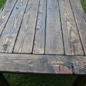 May include: A rectangular wooden table featuring a dark stained finish with visible wood grain patterns and knots. The tabletop is constructed from several parallel planks joined together, framed by perpendicular boards at each end.