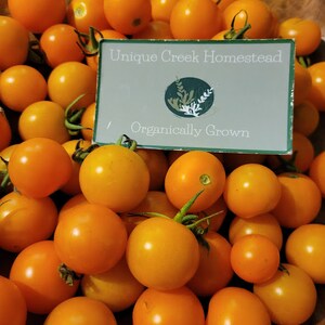 Pu&ograve; includere: Una ciotola di legno piena di pomodorini arancioni brillanti. Un cartello al centro recita "Unique Creek Homestead" e "Organically Grown". I pomodori hanno gambi verdi e un colore vibrante e fresco.