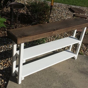 May include: A white wooden console table with two shelves and a dark brown wood top. The table is standing on a gravel surface.