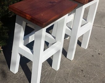 May include: Two white wooden stools with brown wooden tops. The stools are simple in design with a square top and four legs.