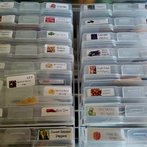 May include: A clear plastic storage container with multiple drawers, each containing small packets of seeds. The drawers are labeled with the names of different pepper varieties, including Sweet Banana Peppers, Habanero Peppers, and Cayenne Peppers.