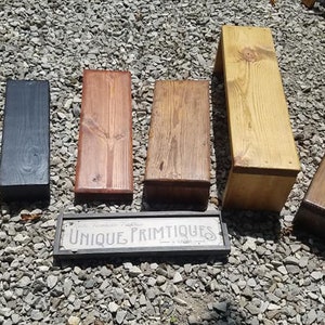 May include: Six wooden shelf brackets in various wood stains, including natural, black, dark brown, and light brown. The brackets are arranged in a row on a gravel surface. A sign reads "Unique Primitives".