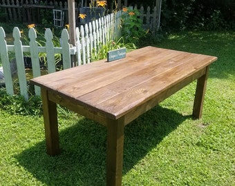 May include: A rustic wooden table with a dark brown stain. The table is made of reclaimed wood and has a simple, farmhouse style. The table is sitting on a grassy lawn.