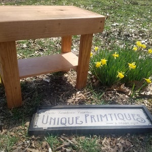 May include: A small, handcrafted wooden side table with a lower shelf. The table is made of light-colored wood and has a simple, rustic design. A sign that reads "UNIQUE PRIMTIQUES & DÉCOR" is in front of the table. Yellow daffodils are in the background.