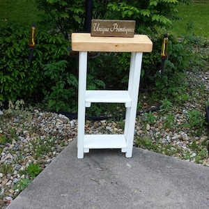 May include: A small, white wooden plant stand with two shelves and a natural wood top. The sign on top reads "Unique Primitives". The stand is outdoors, set on a concrete surface, with greenery in the background.