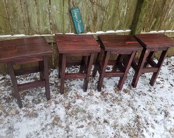 May include: Four dark brown wooden stools with a simple design. The stools are arranged in a row on a snowy surface. The text "Unique Antiques" is visible on a sign in the background.