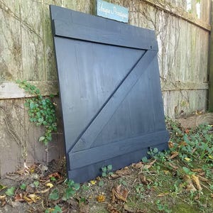 May include: A black wooden door with a diagonal brace and a single horizontal brace. The door is leaning against a wooden fence with vines growing on it. The sign "Unique Printables" is visible above the door.