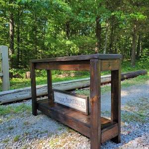 May include: A dark stained wooden console table with a lower shelf and four sturdy legs. A rectangular sign with the text "UNIQUE PRIMTIQUES" is displayed on the lower shelf. The table is outdoors, set against a backdrop of trees and gravel.