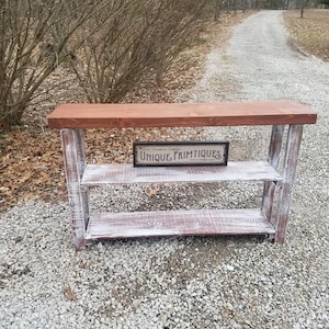 May include: Whitewashed wooden console table with a brown stained top and two shelves. The table has a rustic farmhouse style and features a sign that reads "Unique Primitives".