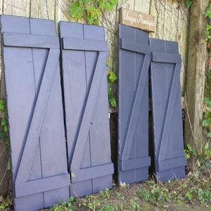 May include: Four dark blue wooden shutters with a diagonal design. The shutters are leaning against a wooden fence with vines growing on it.