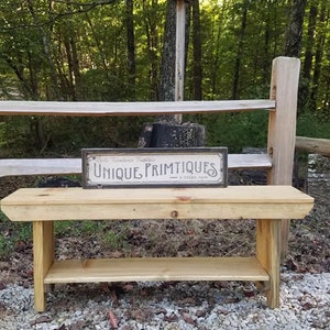 May include: A rustic wooden bench with a shelf, painted in a natural wood color. The bench is sitting on a gravel path in a wooded area. A sign above the bench reads "Unique Primitives".