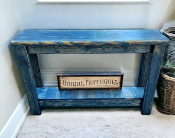 May include: A blue wooden console table with a distressed finish and a single shelf. The table has a sign on the shelf that reads "Unique Primitives".