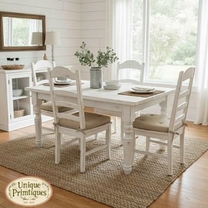 May include: A white wooden dining table with four matching chairs featuring beige seat cushions. The table is set with plates and a green plant in a metal vase. A large rectangular rug sits underneath the furniture set in a bright room.