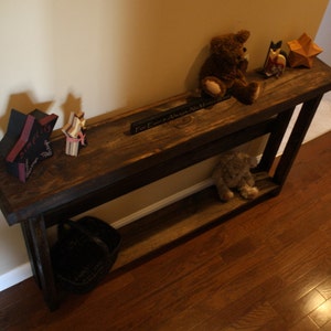 May include: A dark brown wooden console table with a shelf. The table has a distressed finish and is decorated with a variety of items, including a star, a teddy bear, and a sign that reads "I'm Home, Always Welcome".