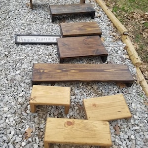 May include: A collection of eight wooden risers in various sizes and finishes. The risers are made of natural wood and have a rustic look. Some of the risers are stained in a dark brown color, while others are left in their natural wood color. The risers are arranged in a staggered pattern on a gravel surface. The text "UNIQUE. PRIMITIQUE." is visible on a sign in the background.