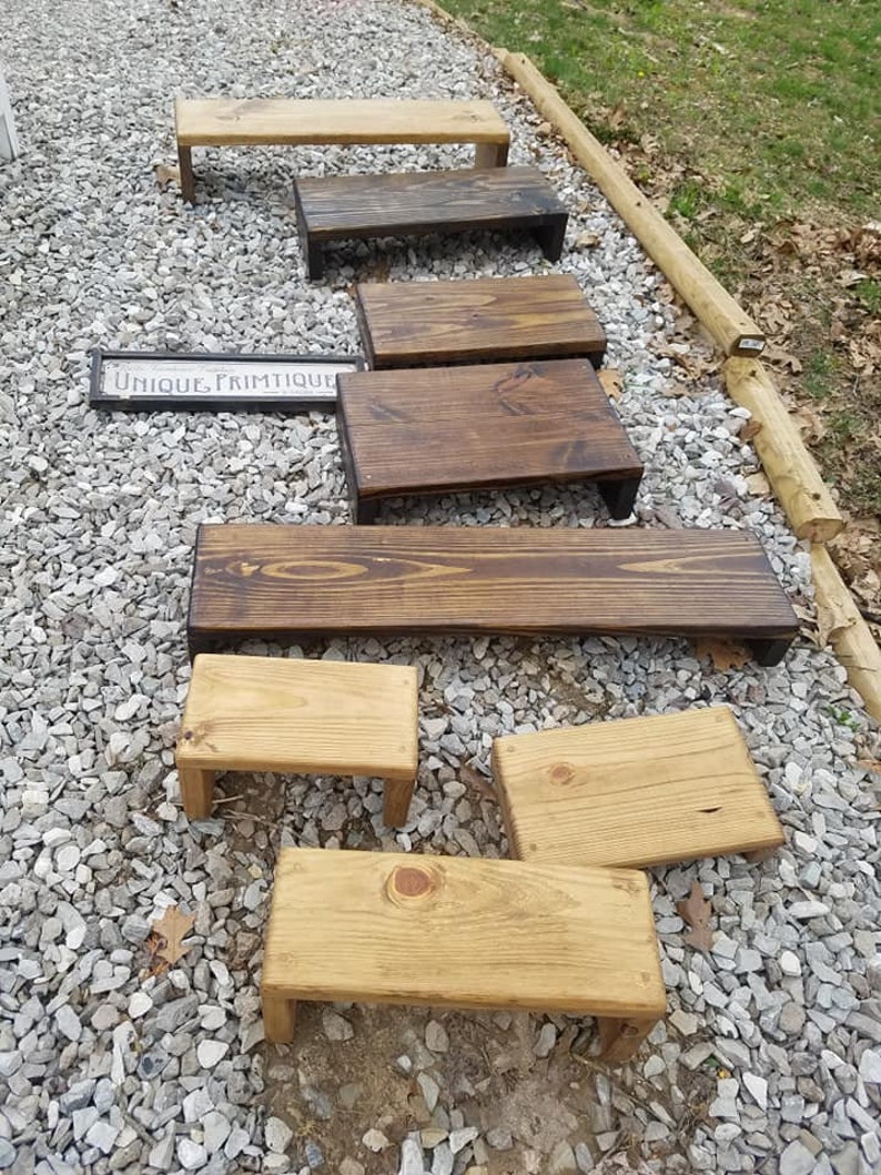 May include: A collection of eight wooden risers in various sizes and finishes. The risers are made of natural wood and have a rustic look. Some of the risers are stained in a dark brown color, while others are left in their natural wood color. The risers are arranged in a staggered pattern on a gravel surface. The text "UNIQUE. PRIMITIQUE." is visible on a sign in the background.