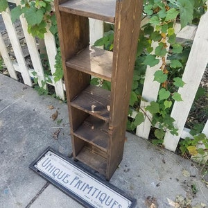 May include: A dark brown wooden shelf unit with five shelves. The shelf unit is standing on a concrete surface. A sign that reads "Cafe Nouveau Primitives & Decor" is lying on the ground.