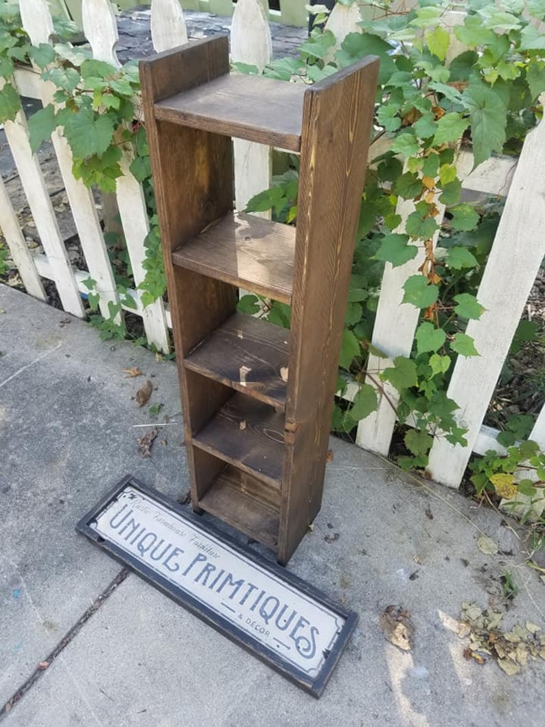 May include: A dark brown wooden shelf unit with five shelves. The shelf unit is standing on a concrete surface. A sign that reads "Cafe Nouveau Primitives & Decor" is lying on the ground.