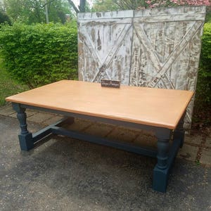 May include: A rectangular wooden table with a smooth, light brown finished top and dark charcoal gray painted legs. The table features four turned legs connected by a horizontal base frame. A small sign on the table surface reads Unique Primtiques.