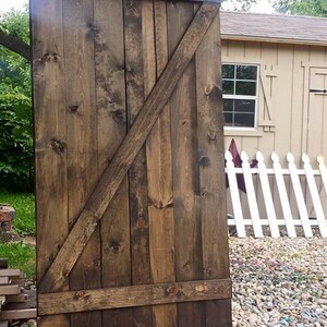 May include: A large, rustic wooden door with a dark brown stain. The door features horizontal planks reinforced with horizontal and diagonal beams. The door is standing upright on a bed of colorful pebbles, with a white picket fence and a shed in the background.