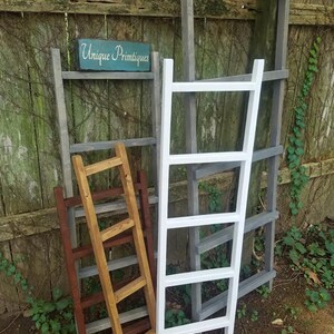 May include: Four wooden ladders leaning against a wooden fence. The ladders are painted in different colors: gray, white, and brown. The sign "Unique Primtiques" is visible on the fence.