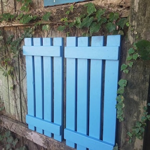 May include: Two light blue wooden shutters with vertical slats are mounted on a weathered wooden fence. A sign above the shutters reads "Unique Primitives" in white script. Green vines and leaves partially cover the fence.