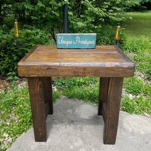 May include: A rustic wooden table with a dark brown stain. The table has a simple design with a rectangular top and four legs. A small teal sign with the text "Unique Primtiques" is placed on the table.