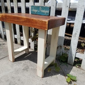 May include: A small wooden table with a distressed white base and a brown top. The table is sitting in front of a white picket fence. A sign that reads "Unique Primitives" is sitting on top of the table.