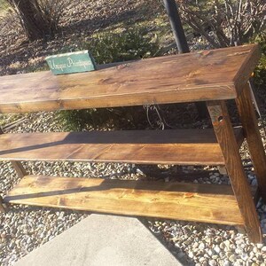 May include: A rustic wooden console table with three shelves. The table is stained a dark brown color and has a distressed finish. The table is sitting on a gravel surface. The sign on the table reads "Unique Boutique".