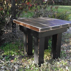 May include: A square wooden stool featuring a dark brown stained top with visible wood grain and four sturdy legs painted in a solid matte black finish. The stool has a low-profile design with a flat surface and thick, block-style legs.