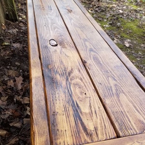 May include: A rustic wooden bench with a warm, brown finish. The bench is constructed from several planks of wood, showcasing the natural grain and knots. The edges are framed, and the bench appears sturdy and ready for outdoor use.