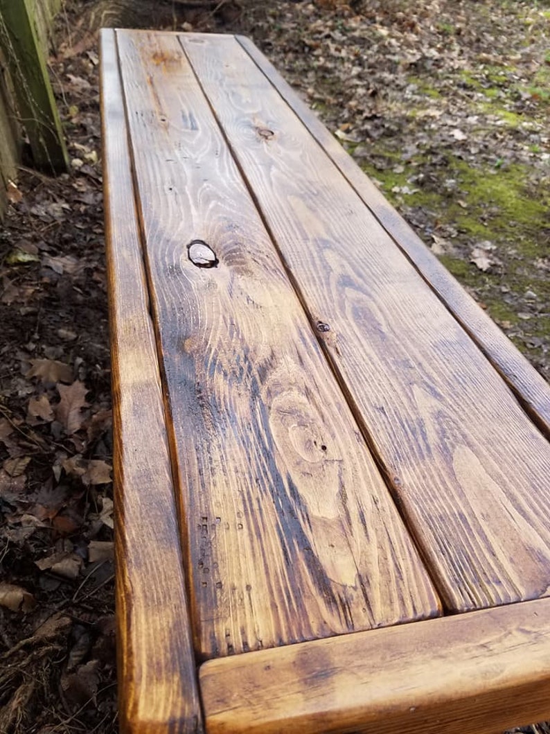 May include: A rustic wooden bench with a warm, brown finish. The bench is constructed from several planks of wood, showcasing the natural grain and knots. The edges are framed, and the bench appears sturdy and ready for outdoor use.