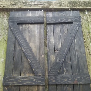 May include: A pair of black wooden shutters with a diagonal design. The shutters are made of reclaimed wood and have a rustic look. The shutters are attached to a wooden fence with the text "Unique Primitives" on a sign above them.