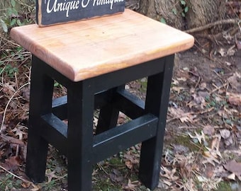 May include: A black wooden stool with a natural wood top. The stool has a simple design with a single shelf. The stool is sitting on a bed of leaves and dirt. A sign that reads "Unique Primtiques" is sitting on top of the stool.