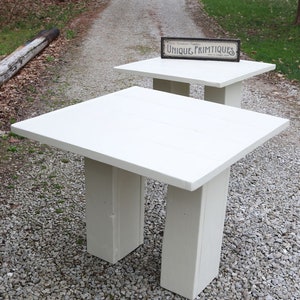 May include: Two white wooden tables with a rustic, farmhouse style. The top table has a sign that reads "Unique Primitives".
