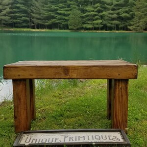 May include: A rustic wooden bench with a rectangular top and four sturdy legs. The bench is made of stained wood and sits on a grassy area near a lake. A framed sign with the words "UNIQUE PRIMTIQUES" is in front of the bench.