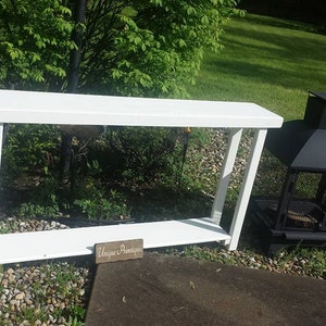 May include: White wooden console table with a single shelf. The table is in a garden setting with a black metal fire pit in the background. A small wooden sign with the text "Unique & Primitive" is in front of the table.