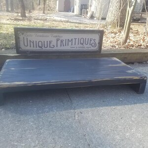 May include: A black wooden riser with a distressed finish. The riser has a cutout on the front and is sitting on a gray concrete surface. A sign above the riser reads "Rustic Farmhouse Furniture Unique Primitives - & decor - "