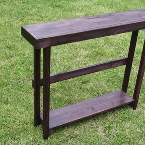 May include: A dark brown wooden console table with two shelves. The table has a simple, rectangular design and is made of solid wood.