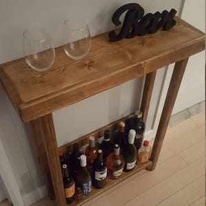 May include: A wooden console table with a brown stain finish. The table has a shelf for storing wine bottles. There are two wine glasses on the top shelf. The word "Bar" is written in black on the top shelf.
