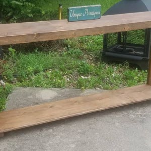 May include: A rustic wooden console table with a brown stain finish. The table has a single shelf and is 60 inches long. The table is sitting on a concrete patio with green grass in the background. A sign that reads "Unique Primitives" is sitting on the table.